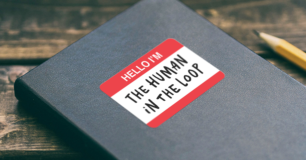 A sticker shaped like a "HELLO I'M" name badge on a black notebook, with a pencil on the dark wooden table beside it. The red-and-white badge is filled in with handwritten capitals reading "THE HUMAN IN THE LOOP".