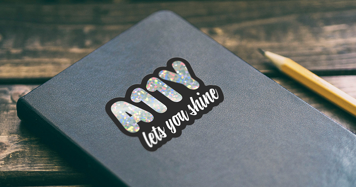 A holographic glitter sticker on a black notebook, next to a pencil on a dark wooden table. Chunky letters spelling "A11Y" sparkle inside a black outline, with "lets you shine" handwritten beneath them.